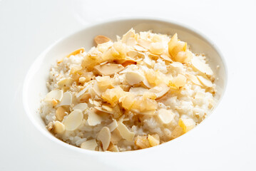 A close-up of a white bowl filled with creamy rice, topped with flaked almonds and pieces of sweetened fruits. The dish is presented on a clean, minimalist white background.