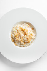 A close-up of a white bowl filled with creamy rice, topped with flaked almonds and pieces of sweetened fruits. The dish is presented on a clean, minimalist white background.