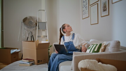 Woman choosing decor online sitting couch with laptop looking on wall new home.