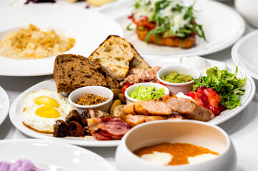 A delicious and hearty breakfast platter featuring fried eggs, sausages, smoked bacon, fresh vegetables, and artisanal bread, accompanied by dips and sauces on a white plate. Perfect for food lovers.