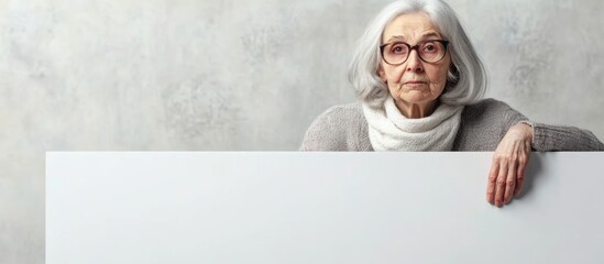 Elderly woman aged 70 to 80 years with gray hair and glasses posing against a neutral gray background holding large empty white sign for text