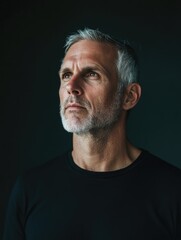 Obraz premium Mature Man with Short Gray Hair and Beard in Black Shirt Posing Thoughtfully Against Dark Background Creating Space for Textual Content