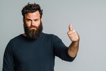 Fototapeta premium Bearded man with a serious expression giving thumbs up gesture against a soft gray background, ideal for conveying support or approval themes.