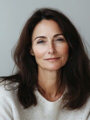 Naklejka premium Elegant portrait of a middle-aged brunette woman with long wavy hair, soft natural lighting, and a neutral gray background ideal for text placement.