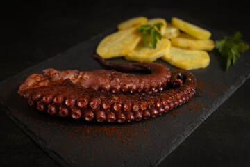 Octopus with potatoes, served on a slate board. Dark background. Octopus legs, grilled octopus, roasted potatoes, octopus with paprika. © mialcas