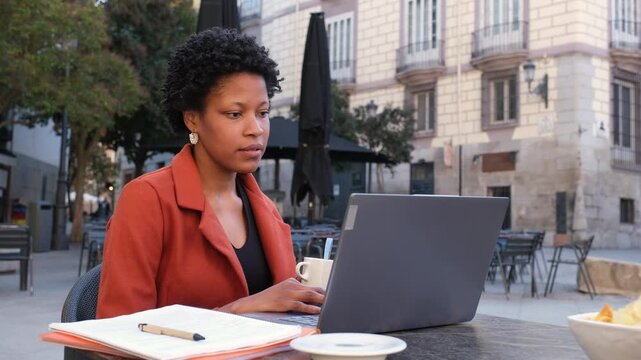 Young black businesswoman working on laptop and drinking coffee at a table in a city square