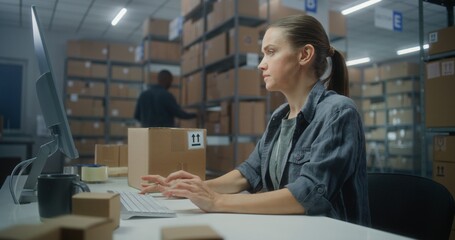 Female warehouse clerk scans parcel with barcode scanner, uses computer, works in postal service. Sorting center employee checks cardboard boxes for shipping to clients. Logistics distribution center.