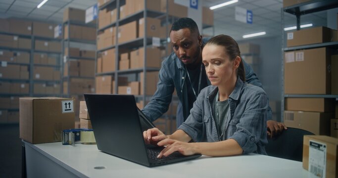 Warehouse or sorting center: Female logistics specialist checks E-commerce online orders on laptop. African American warehouse associate uses tablet computer, consults with colleague. Delivery service