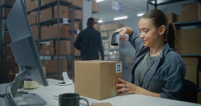 Logistics distribution center: Female post office employee scans cardboard parcel using barcode scanner, works on computer. Sorting center worker checking boxes for delivery. Online shop warehouse.