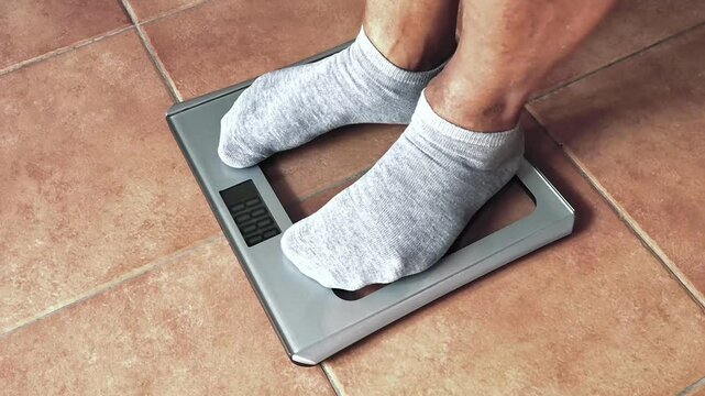 Close-up of a glass weighing scale on which two male feet wearing grey socks stand. 