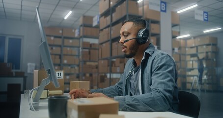 African American logistics coordinator wearing headset chats with customer on call, uses computer,...