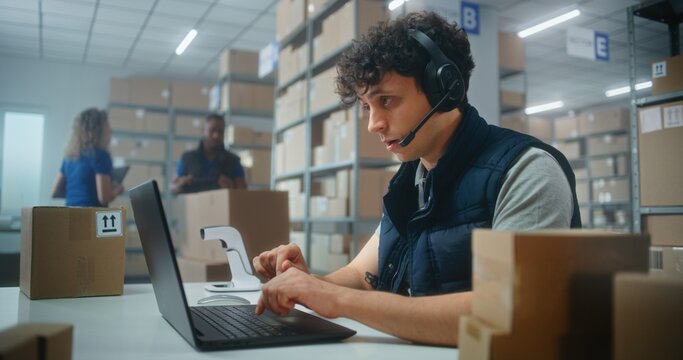 Customer support specialist in logistics wearing headset talks to customer on call, uses laptop computer, works in post office or online store storage. Sorting center workers carrying cardboard boxes.