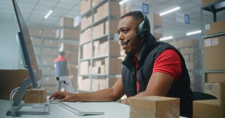 African American customer support specialist in logistics talks on call using headset, uses computer, works in post office or online store storage. Sorting center workers checking cardboard boxes.