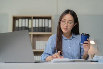 Fototapeta premium Asian businesswoman wearing glasses, working intently on a laptop while sipping coffee at her office desk, balancing professional tasks in a modern corporate environment