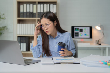 Fototapeta premium Young Asian businesswoman holding a cup of coffee and touching her head with painful expression, suffering from headache, working on a laptop at her workstation