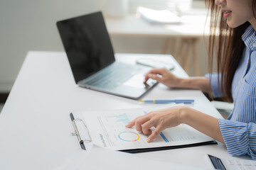 Businesswoman analyzing marketing budget data on a clipboard and using laptop computer, she is working on financial and marketing report in the office