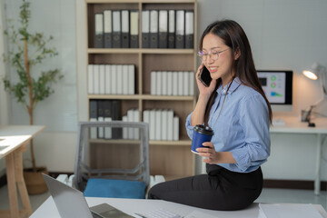 Smiling asian businesswoman is sitting on her desk, talking on the phone and drinking coffee, with a laptop and documents in front of her, in a modern office with a bookcase in the background