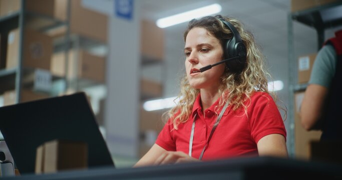 Female customer support specialist in logistics in headset talks to client, uses laptop, works in postal service or online store storage. Sorting center workers checking cardboard boxes. Slow motion. - Powered by Adobe