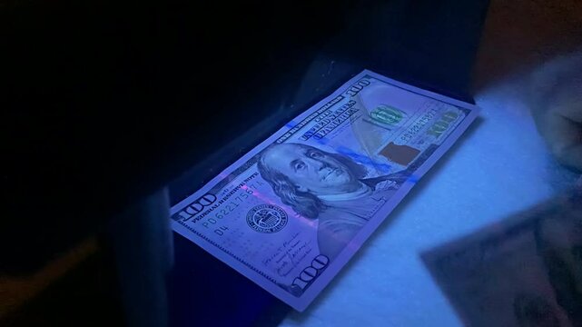 Cashier checking 100 dollar bills under uv light. Bank employee verifying authenticity of one hundred dollar banknotes with ultraviolet lamp.
