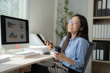 Young office worker is using her smartphone and drinking coffee while looking away and smiling near her computer with graphs and charts on screen