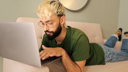 Young blond man with a beard wearing an olive green t-shirt uses a laptop while lying on the sofa in a cozy living room