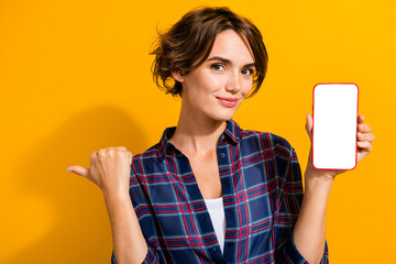 Photo of charming woman dressed plaid shirt show smartphone display indicating at eshop empty space...