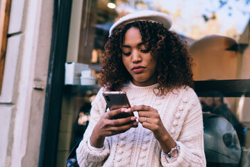 Pensive dark skinned curly woman using smartphone for reading messages and checking notification on smartphone, beautiful african american female influencer watching videos from social networks