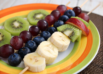  yellow plate with colorful edges containing a skewer of sliced fruits. The fruits include bananas, blueberries, grapes, and kiwi slices. The plate is placed on a table with a polka-dotted tablecloth 