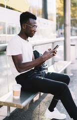 Young dark skinned male traveler in casual wear using app on smartphone for checking public transport schedule, african american man browsing on mobile phone waiting for tram on urban stop