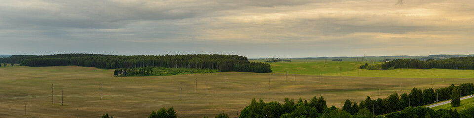 Obraz premium summer agricultural landscape. vast hilly cultivated field with forests, road, poles and distant horizon under cloudy june sky. widescreen panoramic photo in 20x5 format