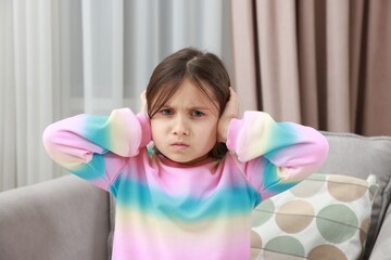 Annoyed little girl covering her ears with pillows from loud sound on armchair at home