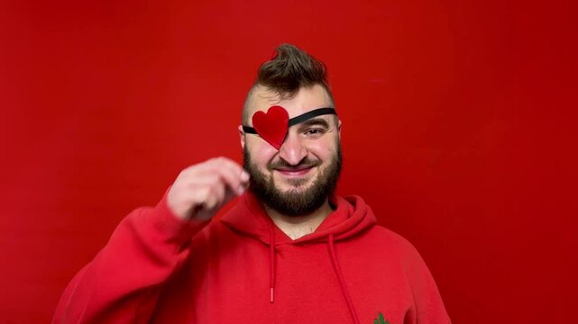 Horizontal 4K video. Young adult white man with beard and mohawk in red hoodie looks at camera, smiles, puts on heart shaped occluder on scarlet background. Concept of Valentine's day, love