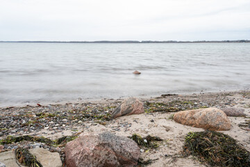 Eine Langzeitbelichtung der Ostsee, das Wasser bewegt sich nur leicht, ein Stein schaut aus der See heraus. Am Strand sieht es Natur belassen aus auf der Gegenüber liegenden Seite ein Küstenstreifen. 