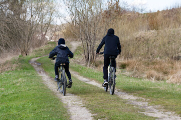 Naklejka premium Two people on bicycles ride along a dirt path through a grassy field. Trees and shrubs line the path. A peaceful outdoor scene.