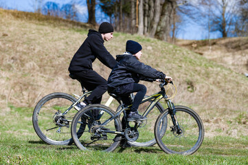 Fototapeta premium Two young people are biking together, enjoying a sunny day outdoors. The path winds through a grassy landscape. They are wearing dark clothing.