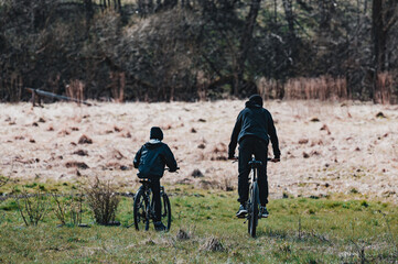 Fototapeta premium Two figures on bicycles ride across a grassy field, enjoying a peaceful outdoor adventure. The background features a blurred woodland and dry grass.
