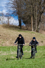 Fototapeta premium A picturesque scene of two young boys cycling across a vibrant green field under a clear blue sky. The backdrop includes a line of trees and a gently sloping hill.