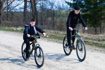 Naklejka premium Two young boys cycle along a country lane. Sunlight illuminates the scene as they enjoy an outdoor adventure. The path is dirt, and trees line the roadside.