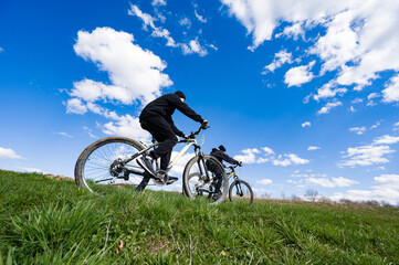 Naklejka premium A low-angle shot of two individuals cycling uphill on mountain bikes. The vibrant blue sky and fluffy white clouds create a beautiful backdrop to their outdoor adventure.