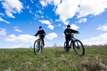 Fototapeta premium Two young cyclists ride mountain bikes on a grassy hill, enjoying a beautiful day with a clear blue sky and fluffy white clouds. A perfect day for outdoor adventure.