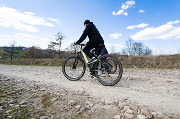 Obraz premium A cyclist rides a mountain bike along a rural dirt road. The sky is bright blue with fluffy clouds. Dry grass and trees line the path.