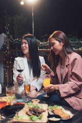 Nighttime Rooftop Rendezvous: Two stylish women share laughter and drinks on a rooftop terrace, enjoying delicious food and good company under the warm glow of evening lights.  