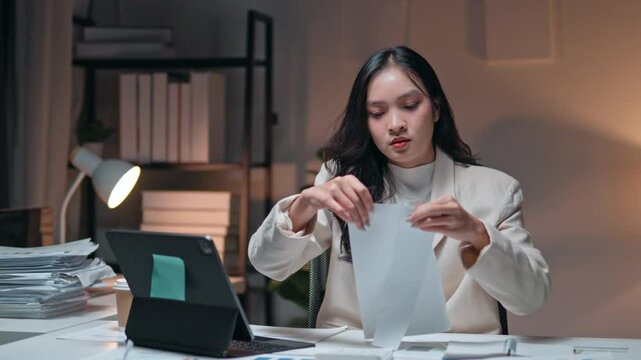 Professional Asian businesswoman ripping important document, experiencing intense workplace stress during late evening hours with dim office lighting
