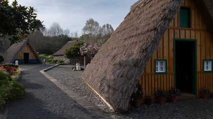 A charming group of traditional A-frame thatched houses with wooden facades, surrounded by cobblestone paths, flowers, and lush greenery.