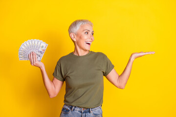 Photo of charming positive woman wear khaki t-shirt holding cash showing arm empty space isolated yellow color background