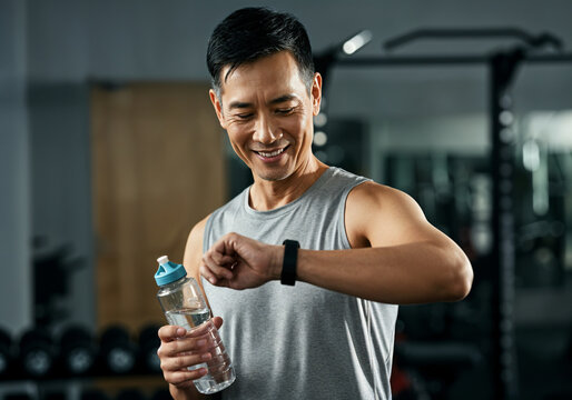 Smiling sportsman checking progress on smartwatch, holding water bottle during workout break at gym