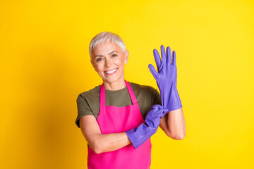 Portrait of pretty aged woman put latex gloves maid pink apron isolated on yellow color background