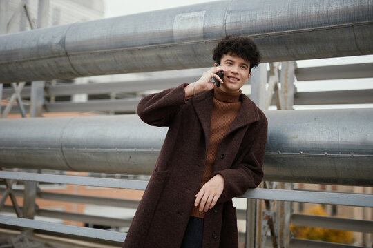 Fashionable man in brown coat talking on phone by metal railing, pipes in background