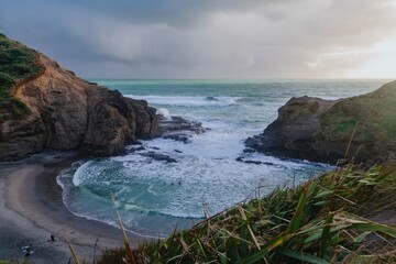 Coastal scene: Rough waves crash against rocky shore; people swim in the ocean. Piha, Auckland, New...