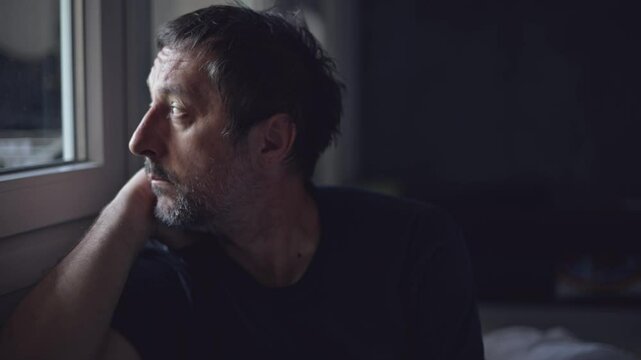 Overthinking and analysis paralysis concept, portrait of pensive unkempt male looking towards the light that is coming from the window, 4K